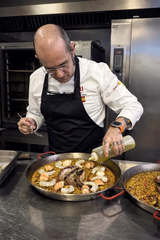 Cristóbal Díaz, con filipina blanca y mandil negro, rocía aceite de oliva sobre una paella de mariscos.